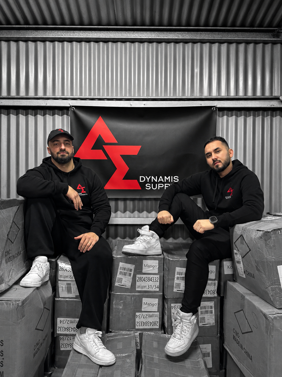Dynamis Supplements founders sitting on stacked boxes with a Dynamis Supplements banner in the background.